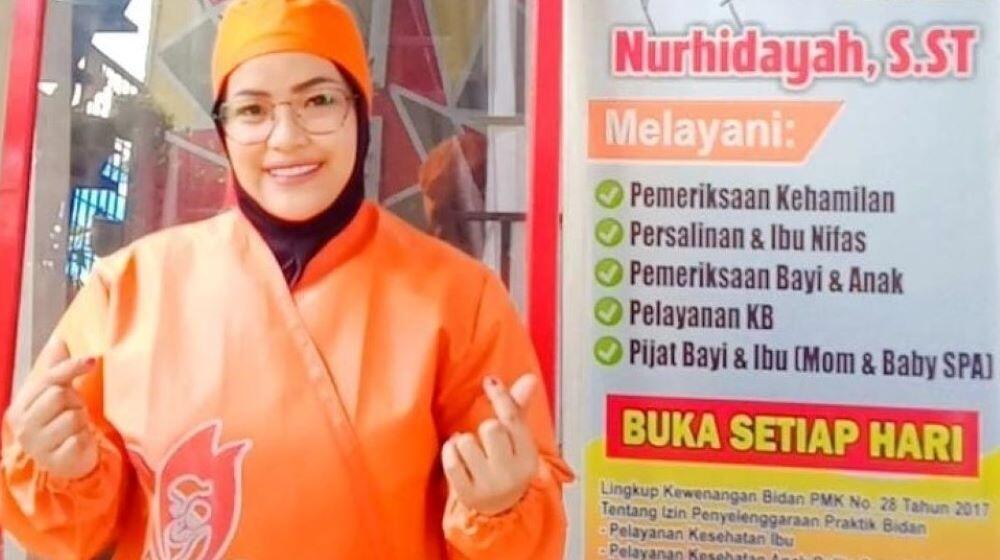 A midwife wearing an orange uniform smiles and poses in front of her private clinic.
